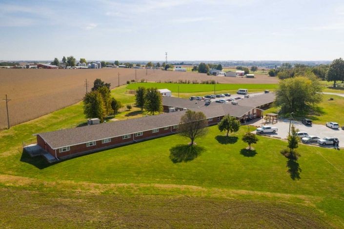 Aerial view of a building with a surrounding parking lot, set in a rural landscape with fields and trees.