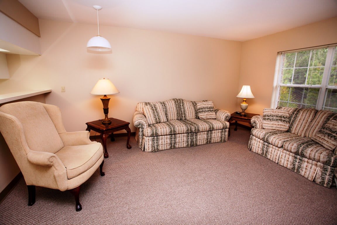 A cozy living room featuring plaid sofas and armchairs, complemented by warm lighting and carpeted flooring.