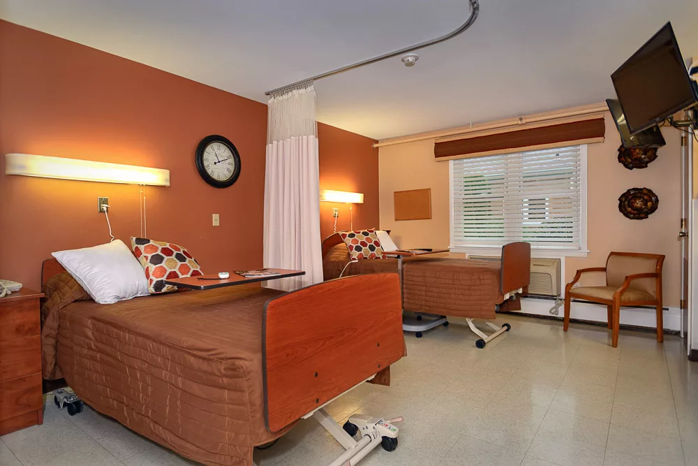 A shared hospital room with two patient beds, featuring warm colored walls and a large window.