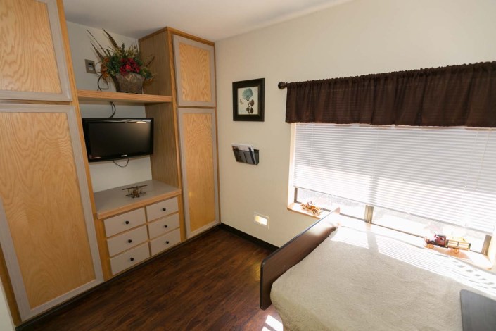 A small bedroom with a wall-mounted television, cabinet, and window letting in daylight.