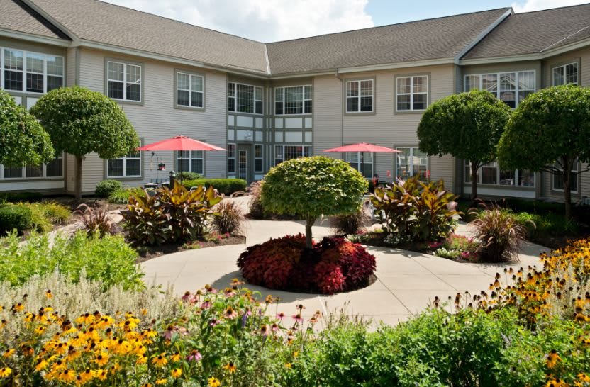 A beautifully landscaped courtyard with colorful flowers, trimmed trees, and sun umbrellas in an outdoor seating area next to a building.