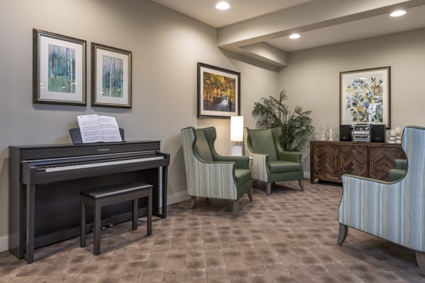 A cozy living room featuring a piano, armchairs, and framed artwork on the walls.