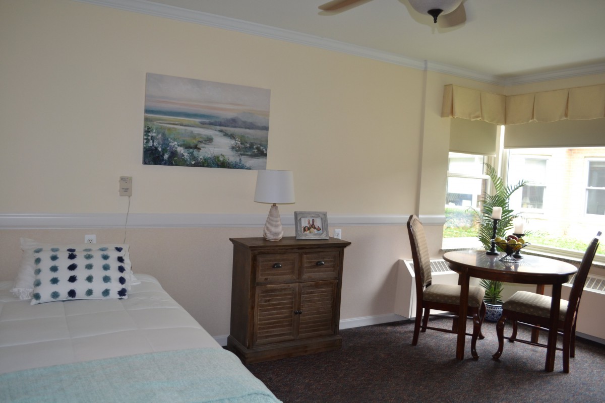 A cozy bedroom with a bed, wooden dresser, and a small table with chairs by the window.