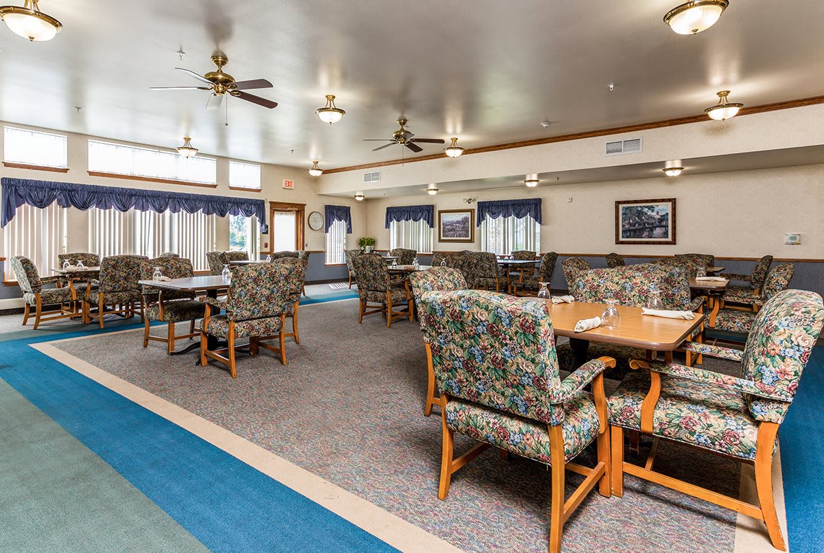 A spacious dining area with multiple tables and chairs, carpet flooring, ceiling fans, and curtained windows.