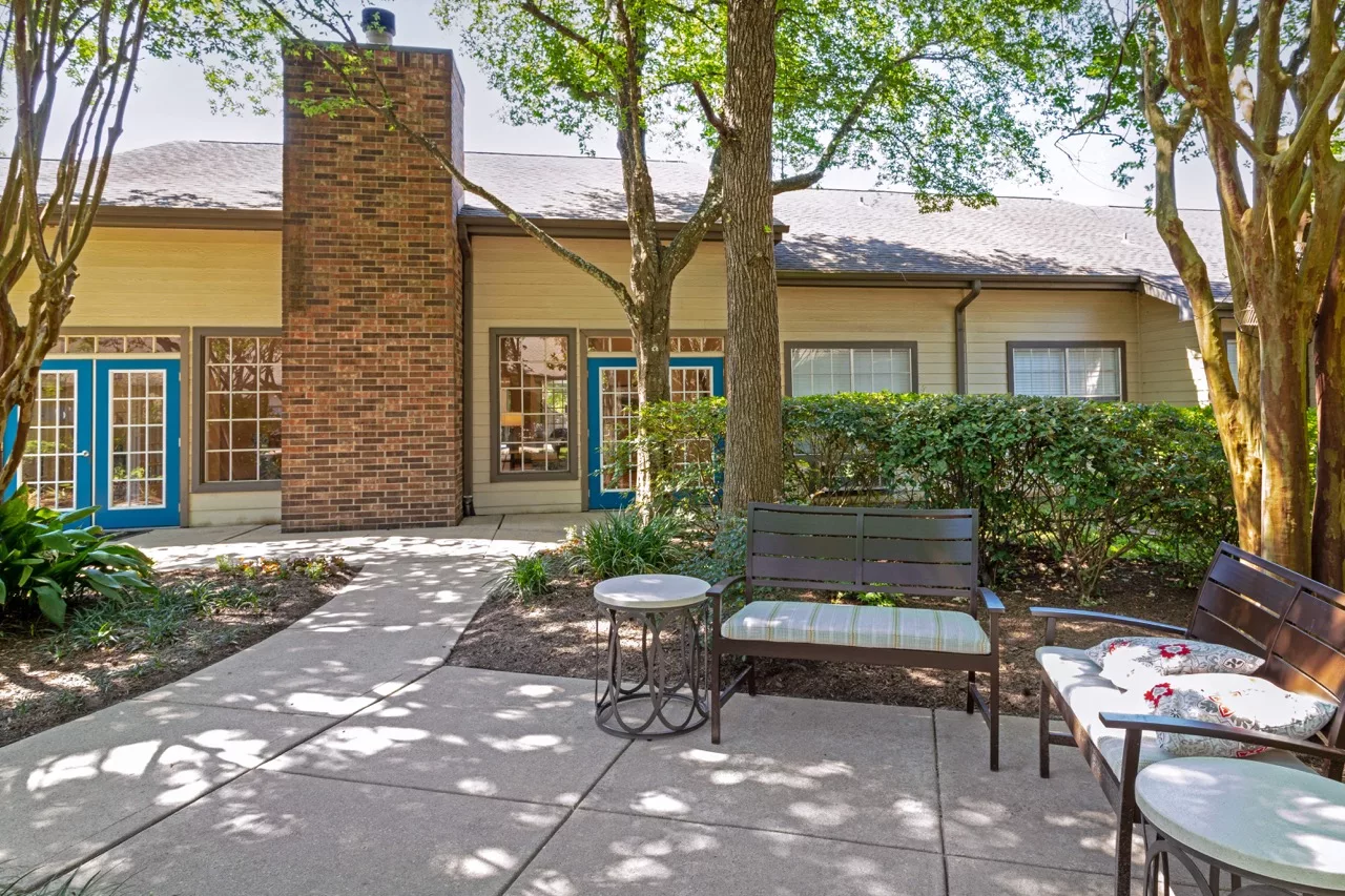 A cozy patio area with a bench and table, surrounded by greenery and trees in front of a house with a brick chimney.