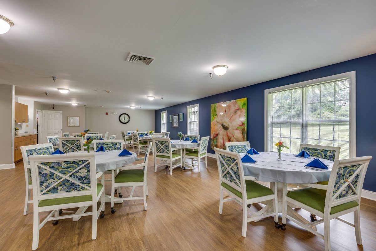 A bright and neatly arranged dining room with multiple tables and chairs, featuring decorative wall art and large windows.