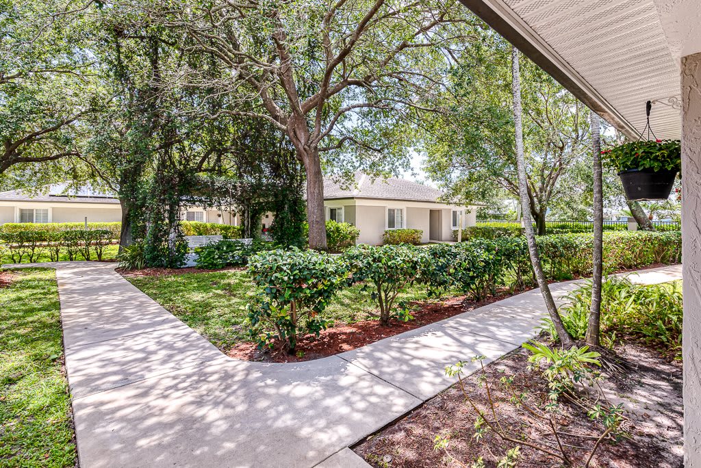A residential house with a landscaped yard, featuring trees and a concrete pathway under sunlight.