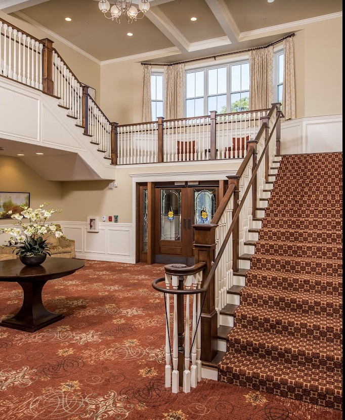 A well-lit foyer featuring a grand carpeted staircase, a central table with floral decoration, and elegant railings.