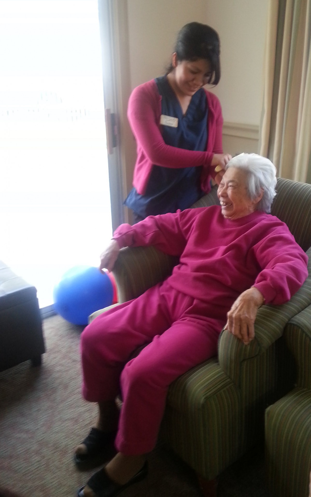 A caregiver assists an elderly woman sitting comfortably in a chair indoors.