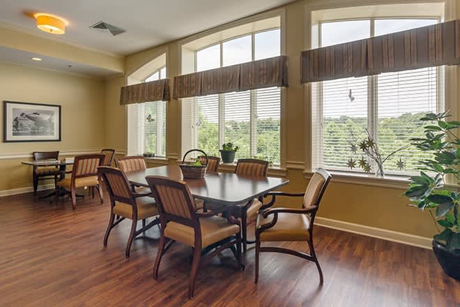 A well-lit dining room with large windows and a wooden table surrounded by chairs.
