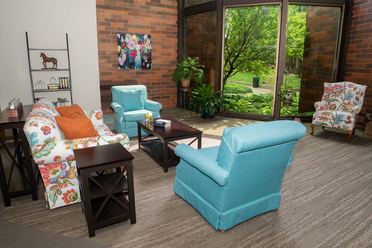 A cozy living room featuring blue armchairs, a floral sofa, and a large window overlooking greenery.