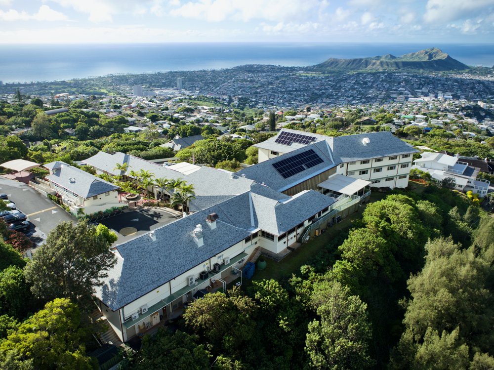 Aerial view of a large building complex surrounded by lush greenery, with a coastal cityscape and ocean in the background.