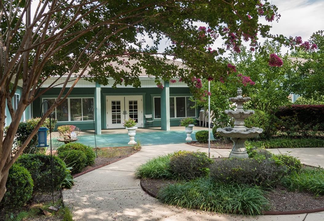 A serene garden pathway leading to a building entrance, surrounded by lush landscaping and featuring a decorative fountain.