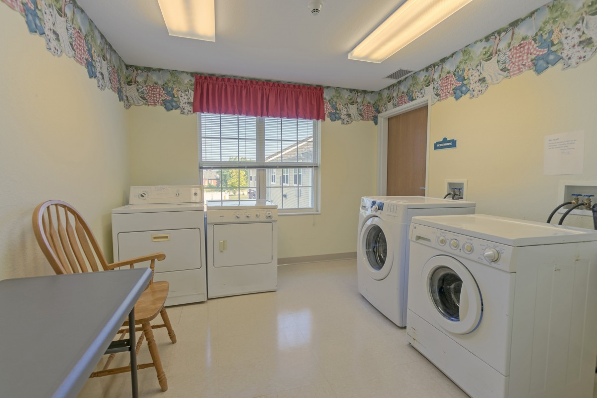 A bright laundry room equipped with washers and dryers, featuring a window with red curtains.