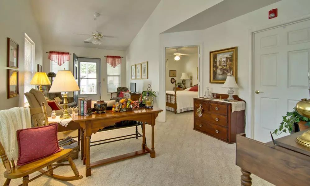 A cozy living room with traditional furniture, featuring a desk and decorations. The room connects to a bedroom, visible through an open doorway.