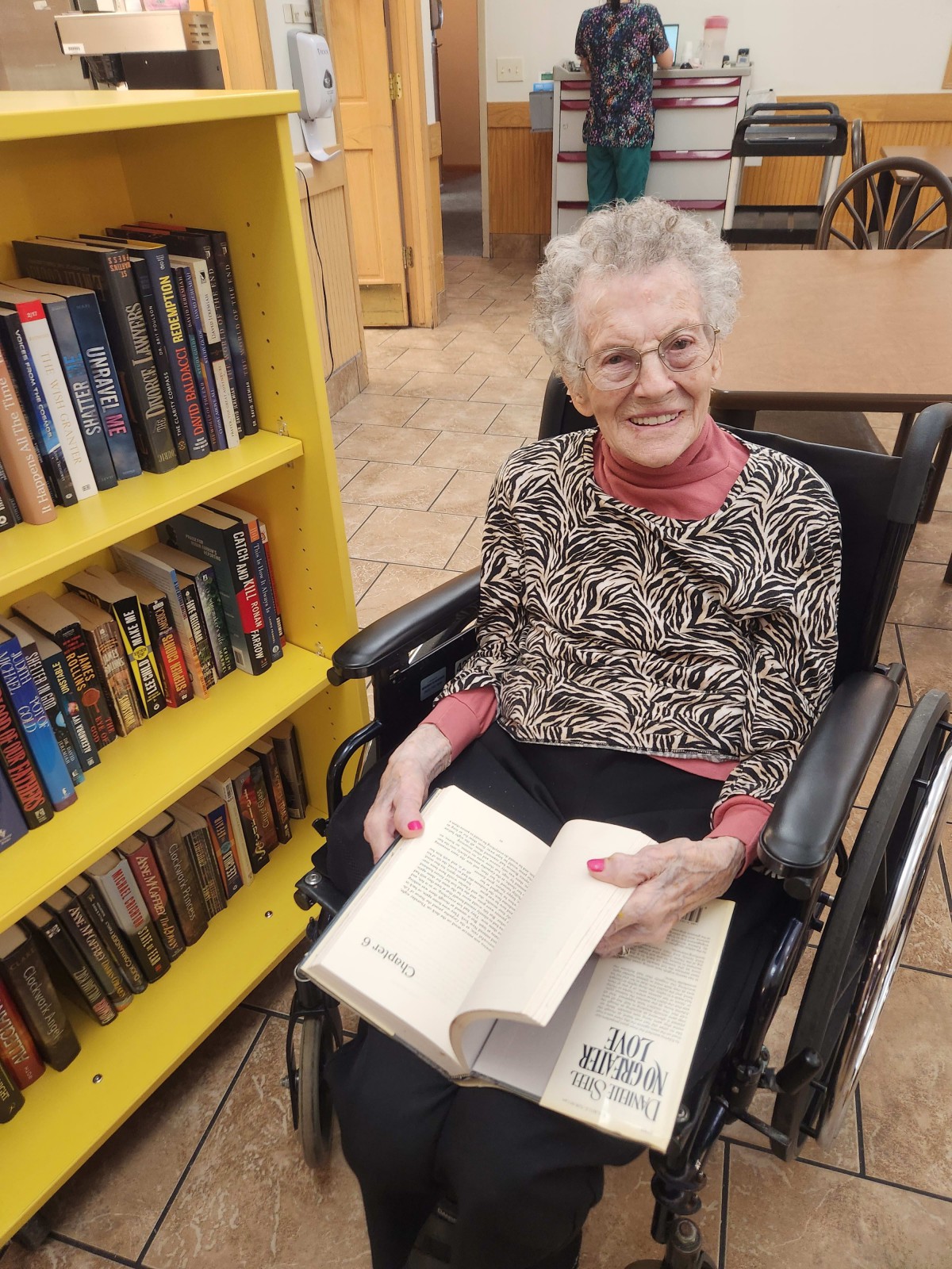 An elderly woman sitting in a wheelchair, enjoying a book in a library setting.