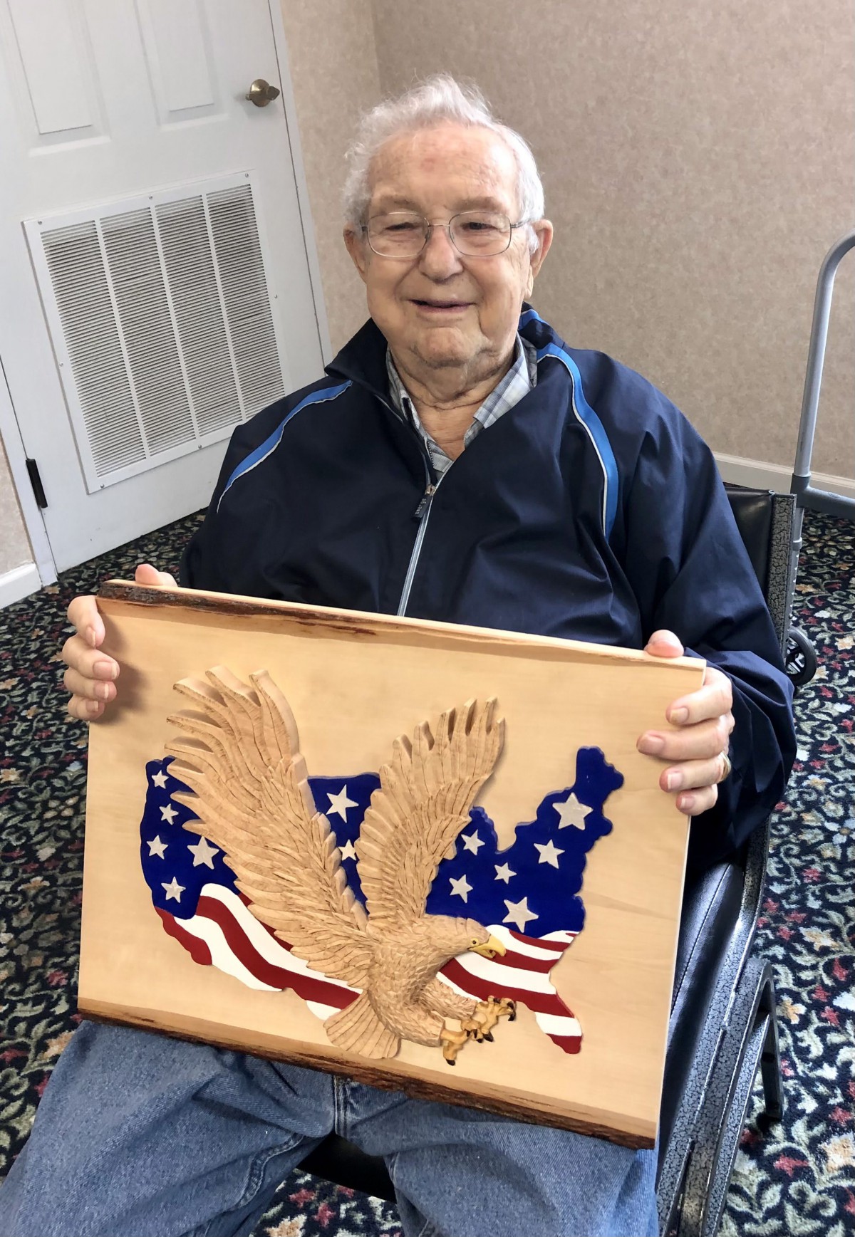 An elderly person in a wheelchair holds a wooden carving featuring an eagle with the American flag.