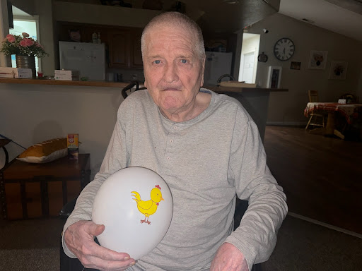 An elderly man smiles while holding a decorative egg featuring a chicken design.