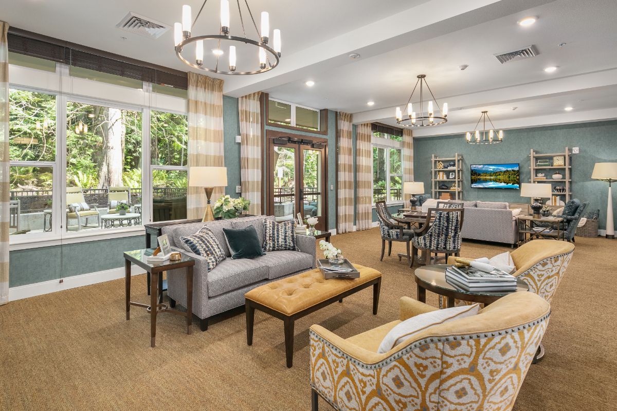 A modern living room with elegant furnishings, featuring a sofa, armchairs, chandeliers, and large windows overlooking a green outdoor view.