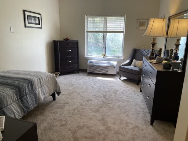 A cozy bedroom with light carpet, featuring a bed, dresser, and a chair by the window.