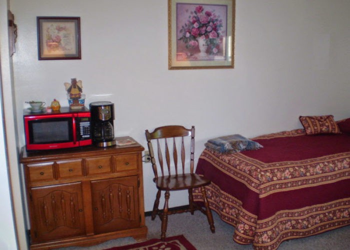 A cozy bedroom featuring a bed with patterned bedding, a wooden chair, and a small table topped with a microwave and coffee maker.