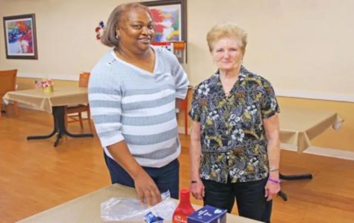 Two women smiling while standing together indoors near a table.