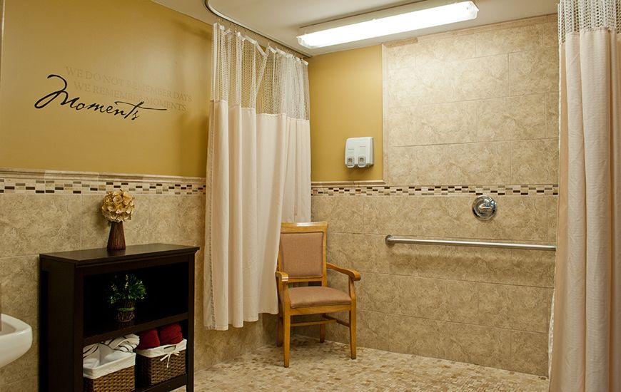 A well-lit bathroom with shower curtain, chair, and handrails on tiled walls.