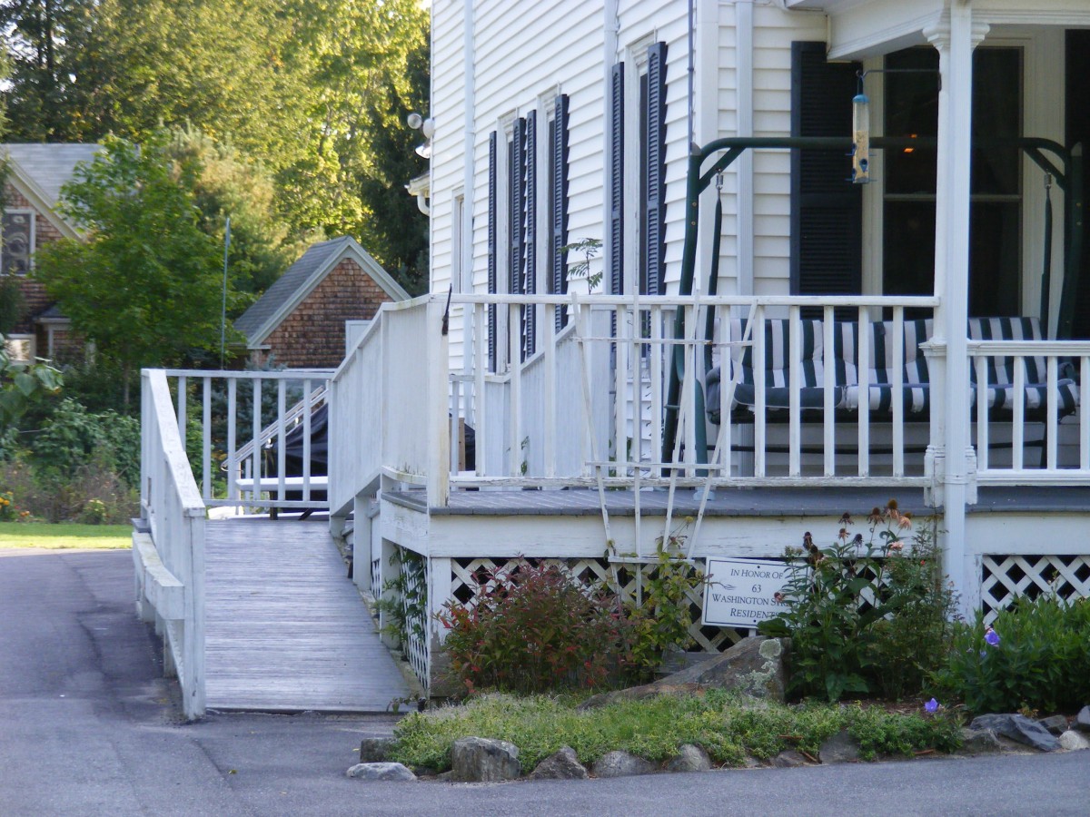 A white house with a front porch, featuring a ramp and a swing, surrounded by greenery.