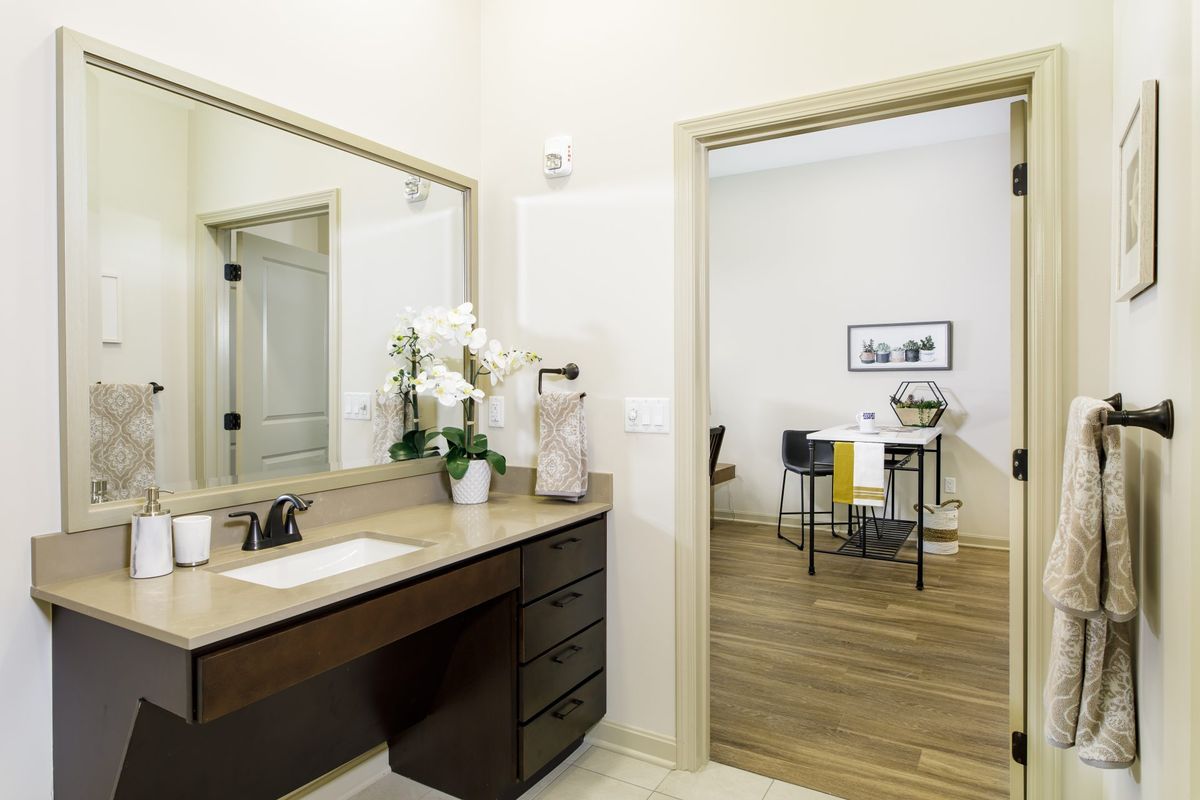 A modern bathroom vanity with a mirror and decorative flowers, leading to an adjacent room with hardwood flooring.