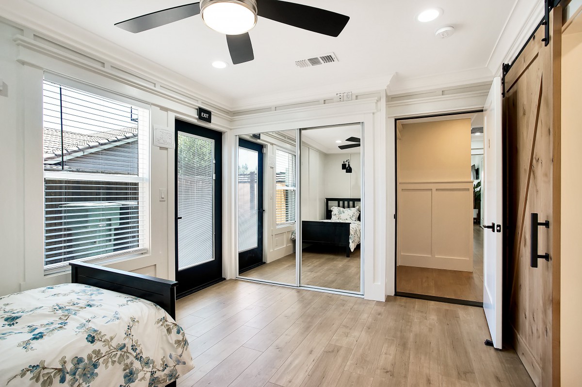 A modern minimalist bedroom featuring wooden floors, mirrored closet doors, and ample natural light.