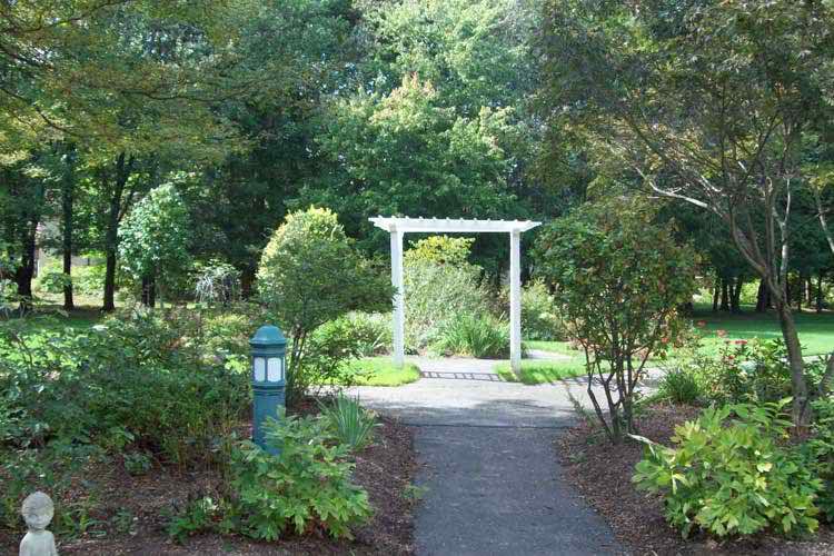 A scenic garden path leading to a white archway surrounded by lush greenery.