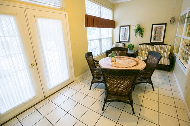 A cozy dining area with a round table and wicker chairs, illuminated by large windows.