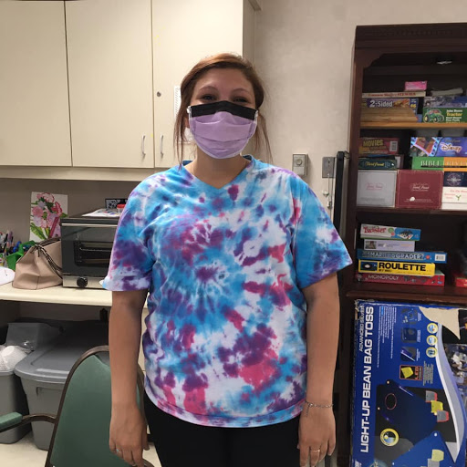 A person wearing a colorful tie-dye shirt and a purple mask stands in an indoor environment.