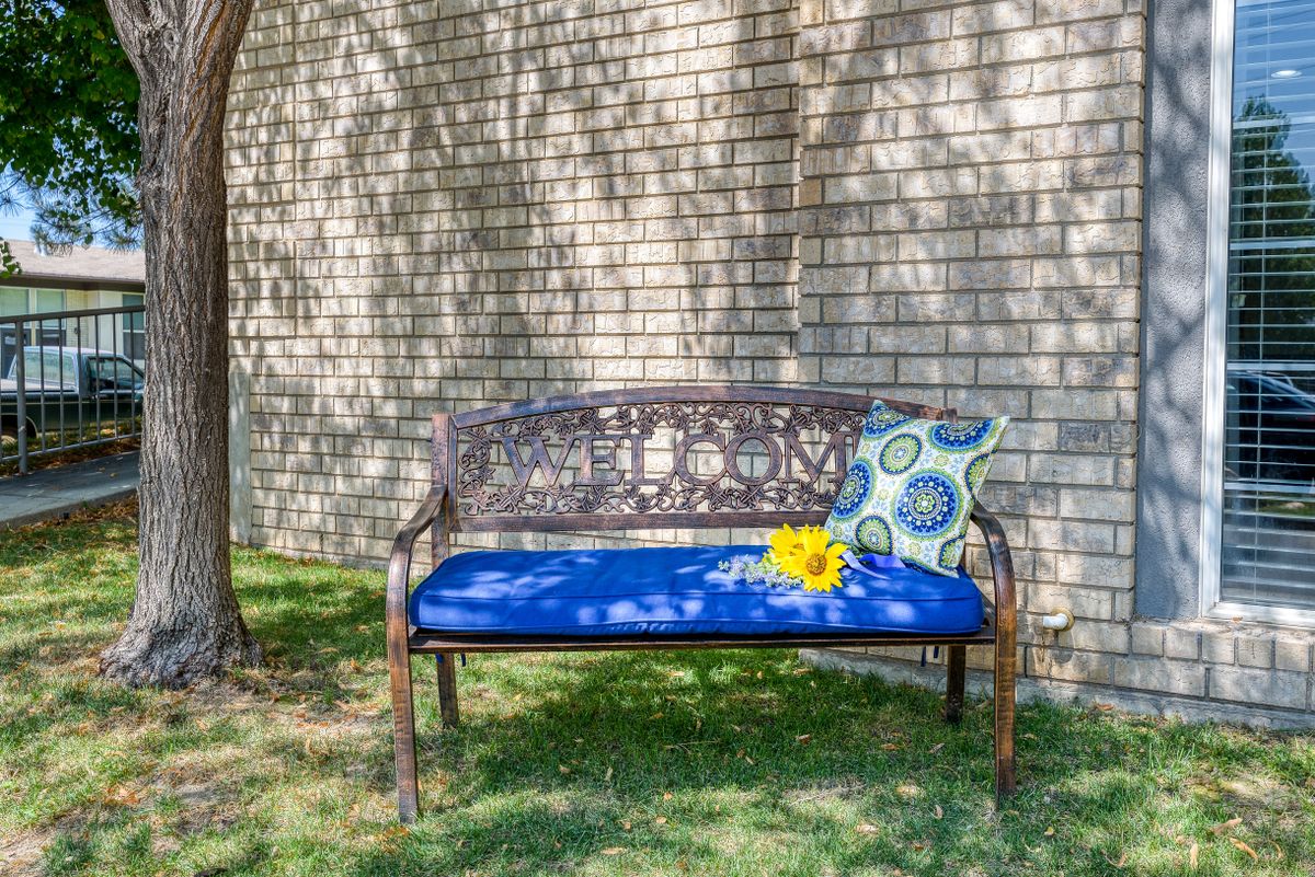 A decorative outdoor bench with a blue cushion, a patterned pillow, and a sunflower resting on it, set against a brick wall with tree shadows.
