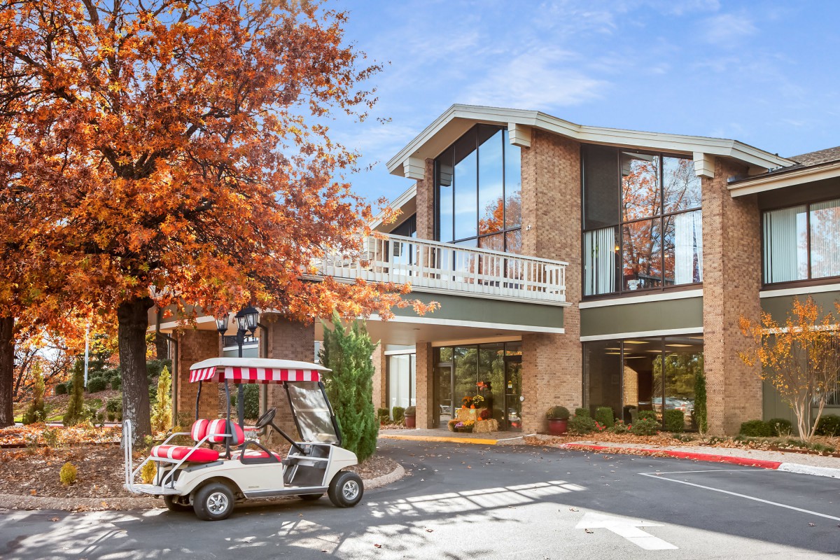 A golf cart parked outside a modern building surrounded by autumn trees.