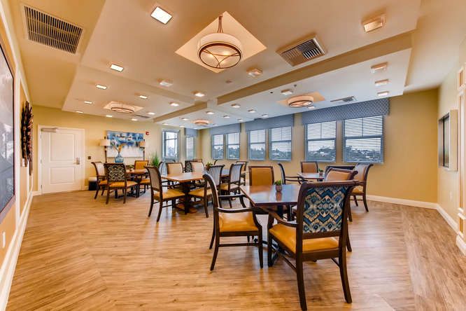 A spacious dining area with multiple tables and chairs, well-lit by ceiling lights and featuring large windows.