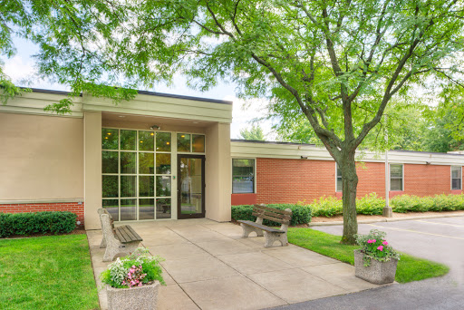 A welcoming entrance to a modern building surrounded by green trees and blooming flowers.