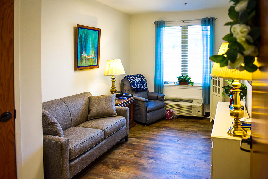 A cozy living room with a sofa, armchair, and a painting on the wall. It features a window with blue curtains, a lamp on the side table, and an indoor plant on the windowsill.