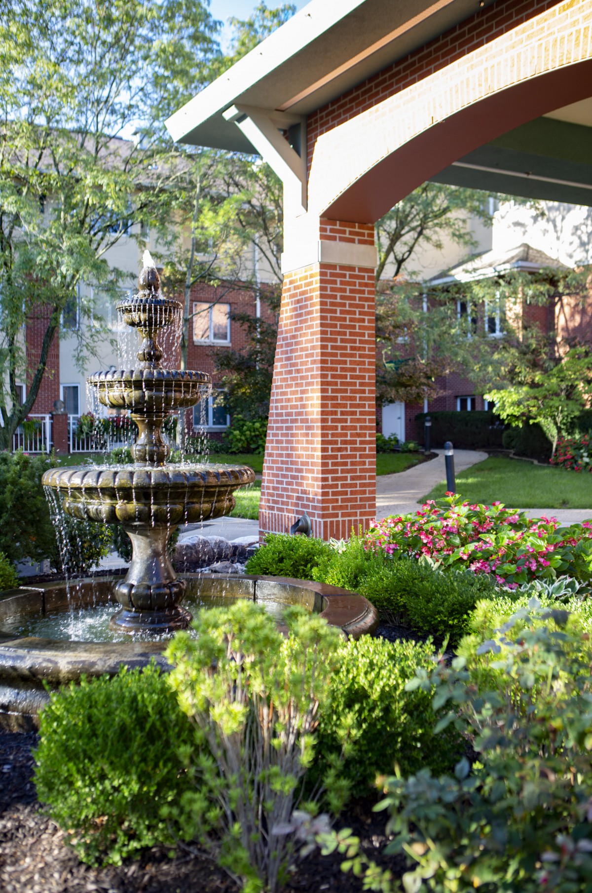 A serene garden featuring a decorative fountain surrounded by lush greenery and vibrant flowers, set against a backdrop of brick architecture.