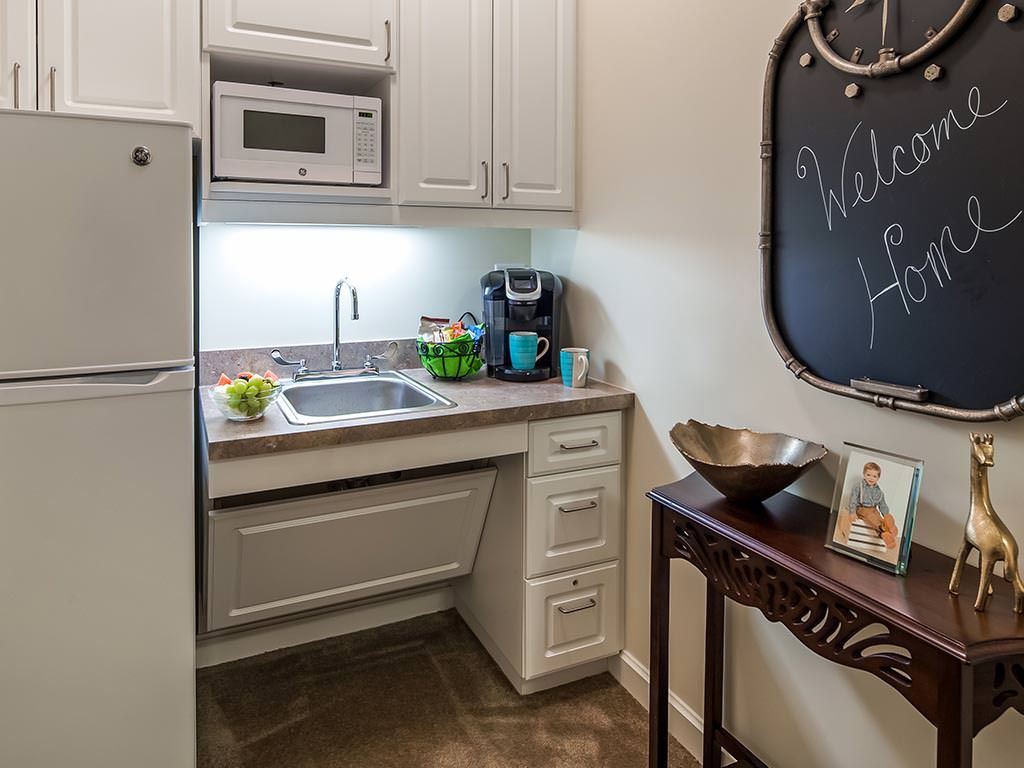 A compact kitchenette with a microwave, sink, and coffee maker, featuring a welcoming chalkboard message.