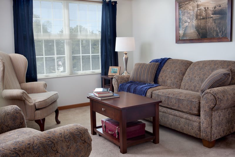 A cozy living room with a sofa, armchair, and coffee table, featuring a lamp by the window with curtains.