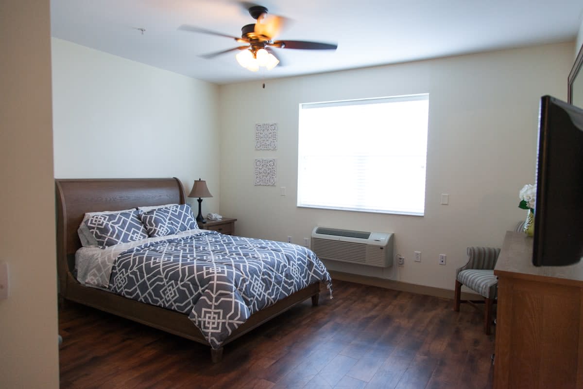 A modern bedroom with a large bed, patterned duvet cover, and hardwood flooring, featuring a chair and lamp near the window.
