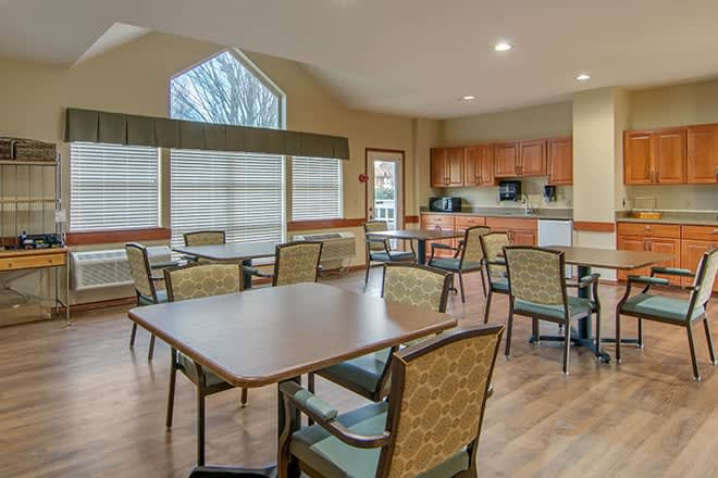 A spacious dining and kitchen area with several tables and chairs, featuring large windows and wooden flooring.