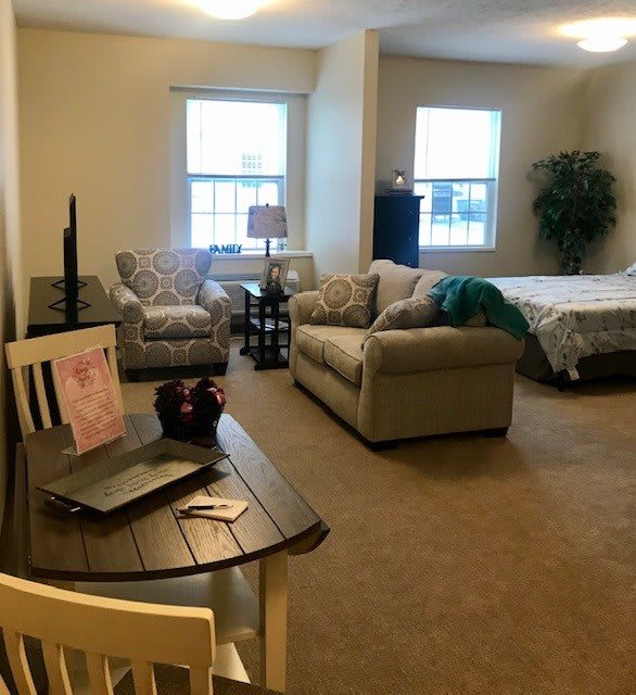 A cozy studio apartment with a combined living and sleeping area, featuring a sofa, bed, small table with a laptop, and large windows.