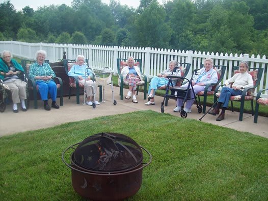 A group of people sitting outside on chairs near a fire pit, with a white fence and greenery in the background.