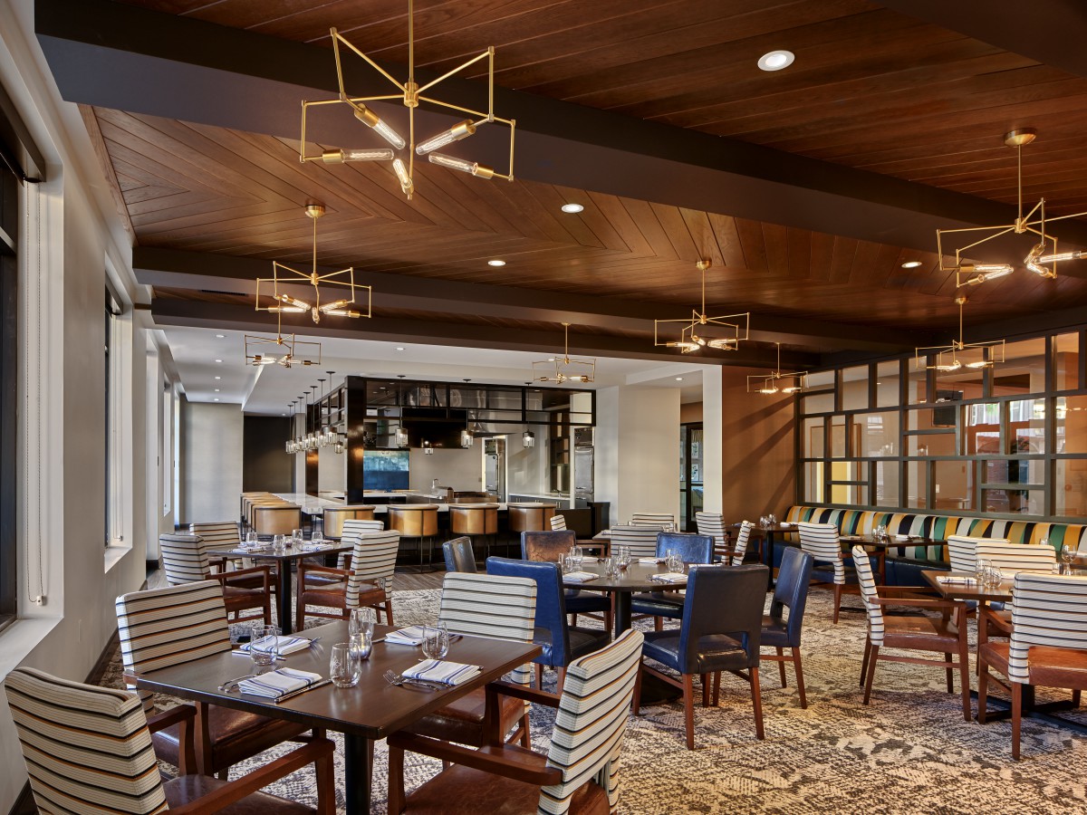A modern restaurant dining area featuring stylish chandeliers, wood paneling, and a well-equipped bar.
