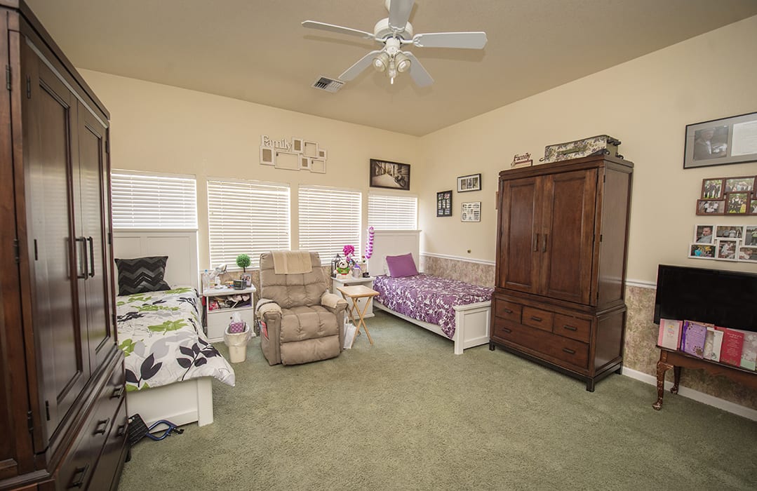 A cozy bedroom with two beds, an armchair, and a wardrobe, featuring a ceiling fan and ample natural light from the windows.