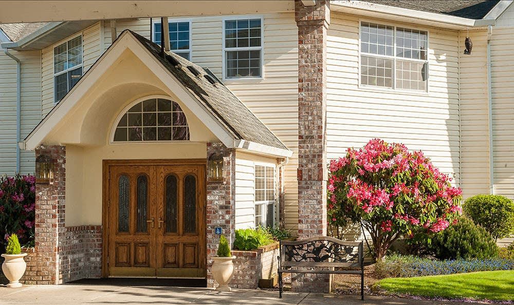 A welcoming house entrance with a wooden double door and well-maintained landscaping, including blossoming flowers.
