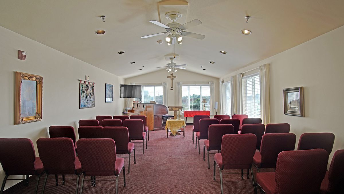 A small chapel with rows of chairs facing a pulpit, featuring ceiling fans and natural light through large windows.