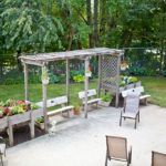 A backyard patio with a wooden pergola and bench seating, surrounded by planters and greenery.
